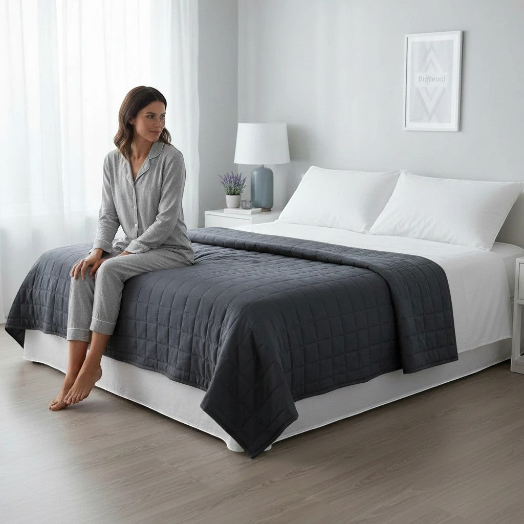 Driftward charcoal grey cotton weighted blanket with quilted pattern on a bed with white bedding. A woman in grey pajamas sits on the edge.